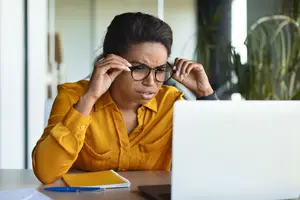 Woman adjusting glasses and looking at a laptop while sitting at a desk in an office.