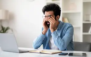 A man sitting at a desk with a laptop and book in front of him, wearing glasses and a blue shirt, he is covering his eyes with his hands.