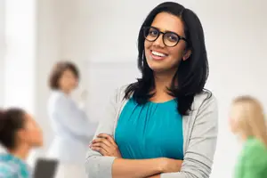 Confident woman wearing glasses and a teal top standing in a room with blurred colleagues behind her.