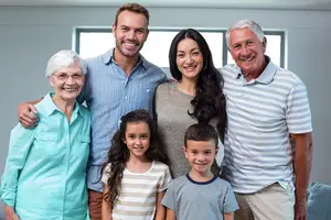 A family of five, including an elderly woman, a man, a woman, and two children, are standing and smiling for a photo in a room with a gray wall and a window.