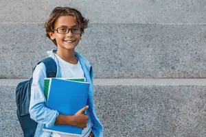 A young boy wearing glasses and a backpack is holding books while standing on the stairs and smiling