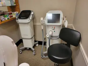Two medical machines with screens are placed in a room with a black chair and a shelf with medical supplies.
