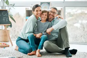 A smiling family of three sitting on the floor in a room with large windows and a blackboard with the letters C and F written on it.