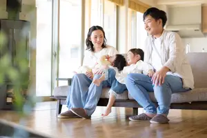 A family of four, including a woman holding a baby, a man, and two children, are sitting on a couch in a living room and smiling for a photo.