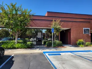 A parking lot with a brick building and some greenery in front of it, a blue sign on the sidewalk, and cars parked on the sides of the building.