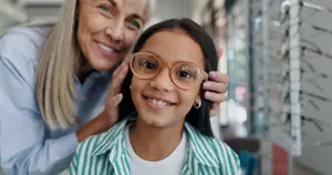 An adult woman adjusting the glasses of a young girl with a smiling face in an optical shop.