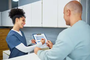 A woman in a blue uniform is sitting at a desk and talking to a man who is holding a clipboard with the text Health Insurance
