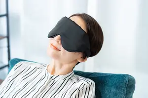 A woman is sitting on a blue couch wearing a black eye mask and a striped shirt.