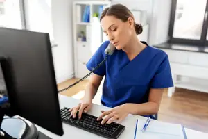 A female medical professional is sitting at a desk and typing on a keyboard while holding a phone to her ear.