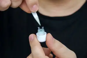 A person is holding a small white object with a transparent cover and a silver tip in their fingers, probably putting a drop of medicine on it.