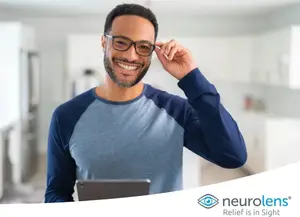 A man with glasses smiling in a room with a tablet in his hand, the brand name Neurolens is on the bottom right.