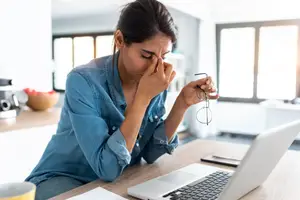 A woman adjusting her glasses while sitting in a room with a laptop on a table