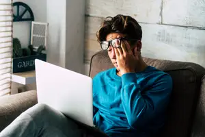 Man sitting on a couch with a laptop rubbing his eyes