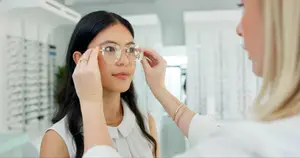 A woman is trying on glasses at an optical store while another woman helps her adjust them in front of a mirror.