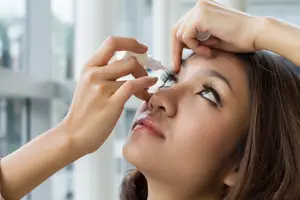 A woman applying eye drops while looking at the mirror in a room with glass windows