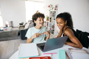 Two people are sitting at a table, a young boy is laughing while holding a pencil, a woman is looking at a laptop, and there are books and other items on the table.