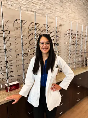 A smiling woman in a white lab coat stands in front of a wall displaying various eyeglasses.