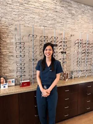 A smiling woman in blue scrubs stands in front of a wall with glasses on display at an eye clinic.