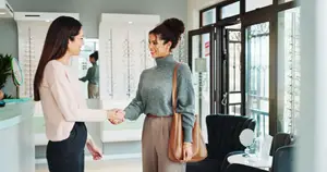 Two women shaking hands in a room with a counter, chair, and mirror. One woman is wearing glasses and the other is holding a handbag.