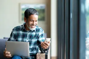 A man with a beard is sitting on a couch and smiling while looking at his phone and laptop