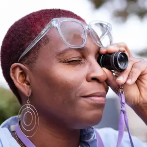 A woman with maroon hair is looking through a telescope with her eyes closed, wearing glasses and earrings