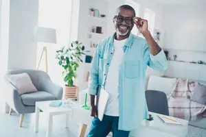 A smiling man holding a tablet and adjusting his glasses in a living room.