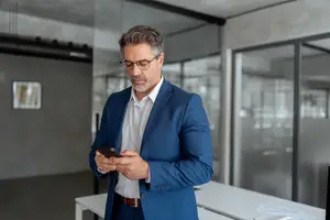 A man in a blue suit is looking at his phone in an office.