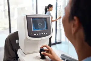 A person operating an ophthalmoscope while a woman stands nearby and another woman looks at the monitor.