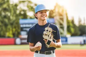 A young man in a baseball uniform holds a baseball and a glove, smiling in a field.