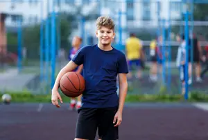 A boy in a blue t-shirt and black shorts holds a basketball on a basketball court with a blue fence and blurry people in the background.