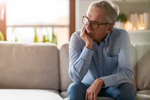 An older man sitting on a couch, looking worried and holding his chin with one hand.