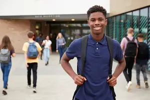 A group of students in a school hallway, including a smiling student in a blue shirt, carrying a backpack, walking towards the school entrance.