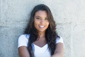 A smiling young woman with long dark hair leans against a gray stone wall and wears a white T-shirt
