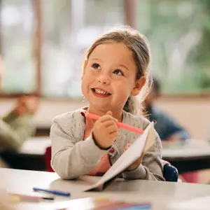 A young girl with blonde hair is smiling and holding a pink pen in her hand while sitting on a chair in a classroom with other students.