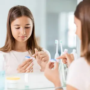A girl with brown hair brushes her teeth in front of a mirror, holding a toothbrush and a bottle of toothpaste.