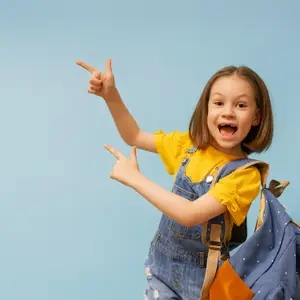 A young girl in a yellow shirt and denim overalls is pointing with her right hand and smiling while carrying a blue backpack with white dots, possibly posing for a photo against a blue background.