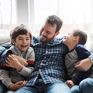 A father with his two sons sitting on a couch in a living room with a bright window in the background