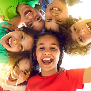 A group of smiling children in a circle wearing green and red shirts, with sunlight shining from above.