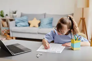 A young girl wearing glasses and a blue blouse is seated at a desk, writing on a piece of paper. A laptop and a pencil stand are on the desk. Behind her, a couch with pillows and a lamp is visible in the background.