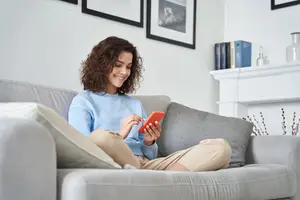 A woman is sitting on a couch, smiling and holding a phone in her hand.