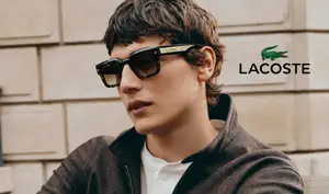 A young man wearing a white shirt, brown jacket, and Lacoste sunglasses stands in front of a brick wall with the Lacoste logo