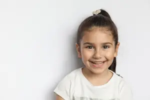 A smiling little girl with her hair tied in a ponytail is standing in front of a white wall wearing a white t-shirt with a silver design