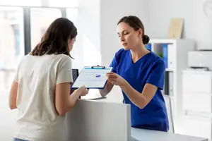 A woman in blue scrubs explains something to another woman in a white shirt at a desk in a clinic.