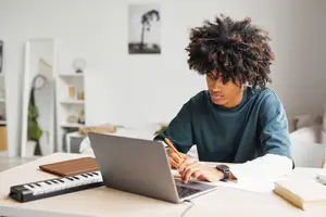 A man is sitting at a table with a laptop, keyboard, and books, looking at the laptop screen.