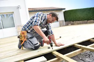 A man wearing a plaid shirt and glasses is working on a wooden floor in a backyard.