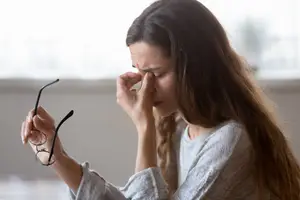 A woman with long hair is holding her glasses and touching her nose with her left hand. She is probably inside a room with a glass window that shows a blurry view of a tree outside.