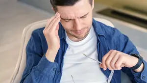 A man is sitting in a chair, holding his head with his left hand, and seems to be adjusting his eyeglasses with his right hand. He is wearing a white t-shirt and a blue jacket. The man is looking down, possibly concentrating on adjusting his eyeglasses. Behind him is a blurry background, which seems to be a room with a glass wall and a white floor.