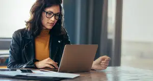 A woman is sitting at a desk, working on a laptop and holding a pen in her right hand. She is wearing glasses and a black jacket. There is a book, a cell phone, a pen, and a notebook on the desk. Behind her is a window with a blue curtain. The light from outside is illuminating the room.