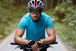 A man wearing a helmet and sunglasses is riding a bicycle on a concrete road with green plants on both sides.