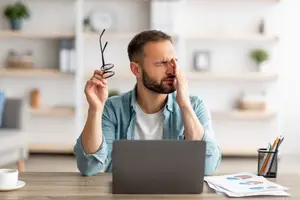 A man adjusting his glasses while sitting at a desk with a laptop and cup.